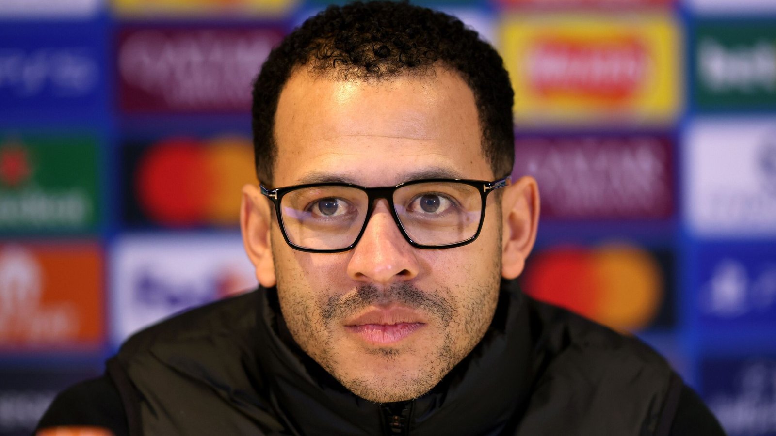 Liam Rosenior, Chelsea-baas, tijdens een persconferentie met een geconcentreerde uitdrukking, een bril dragend tegen een levendige achtergrond van sponsorlogo's.