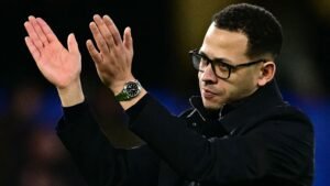 Le manager de Chelsea, Rosenior, applaudissant la foule, portant des lunettes et un manteau noir, avec une montre verte visible à son poignet.