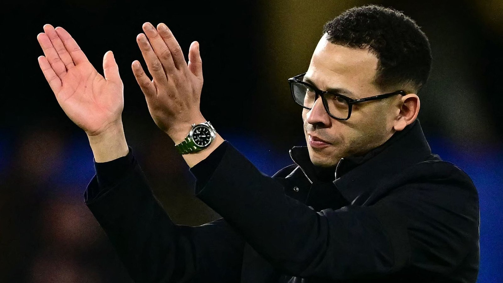 Le manager de Chelsea, Rosenior, applaudissant la foule, portant des lunettes et un manteau noir, avec une montre verte visible à son poignet.