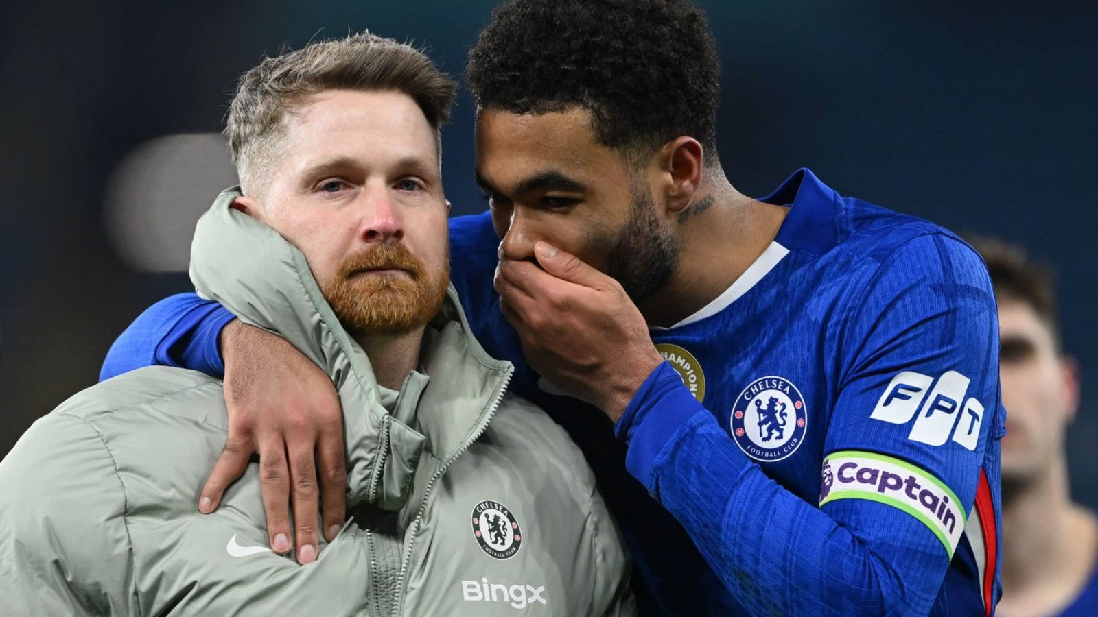 Chelsea-spelers delen een emotioneel moment, waarbij een aanvoerder in het blauw een teamgenoot in een grijze jas omhelst, wat zorg en solidariteit toont.