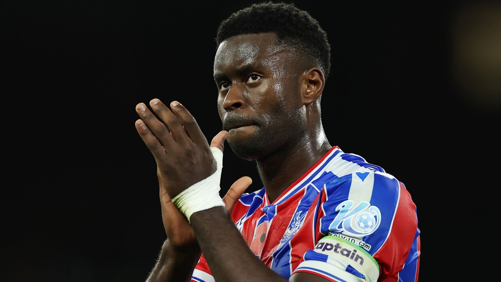 Marc Guehi, in een rood-blauw Crystal Palace-tenue met een aanvoerdersband, gebaart bedachtzaam tijdens een wedstrijd.
