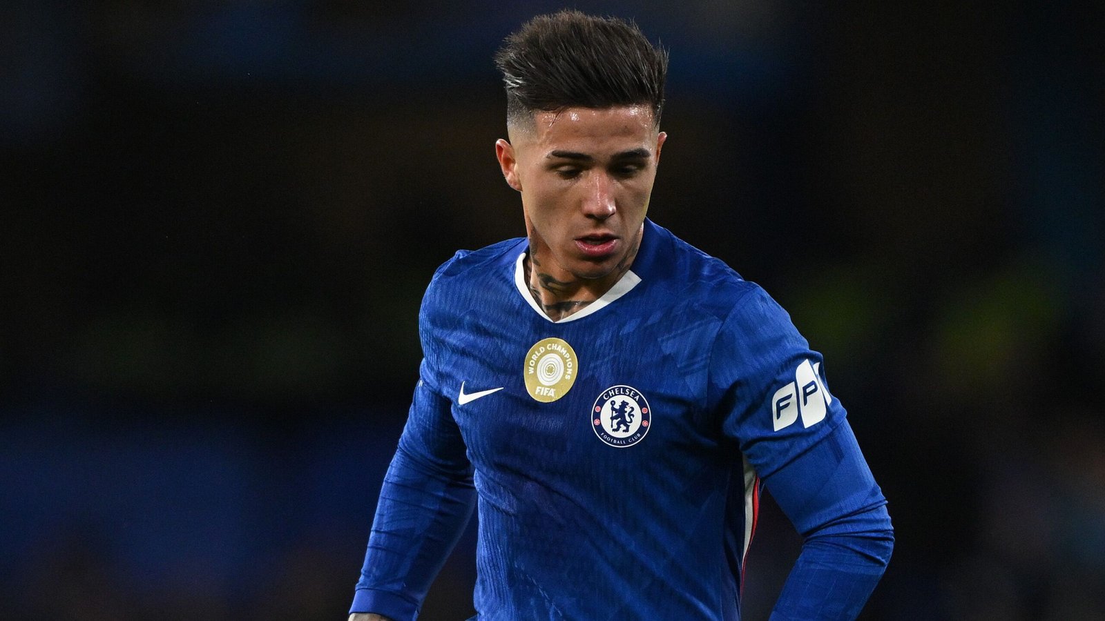 Een Chelsea-speler in een blauw tenue, met een FIFA Wereldkampioenen-insigne, geconcentreerd tijdens een wedstrijd, toont vastberadenheid op het veld.