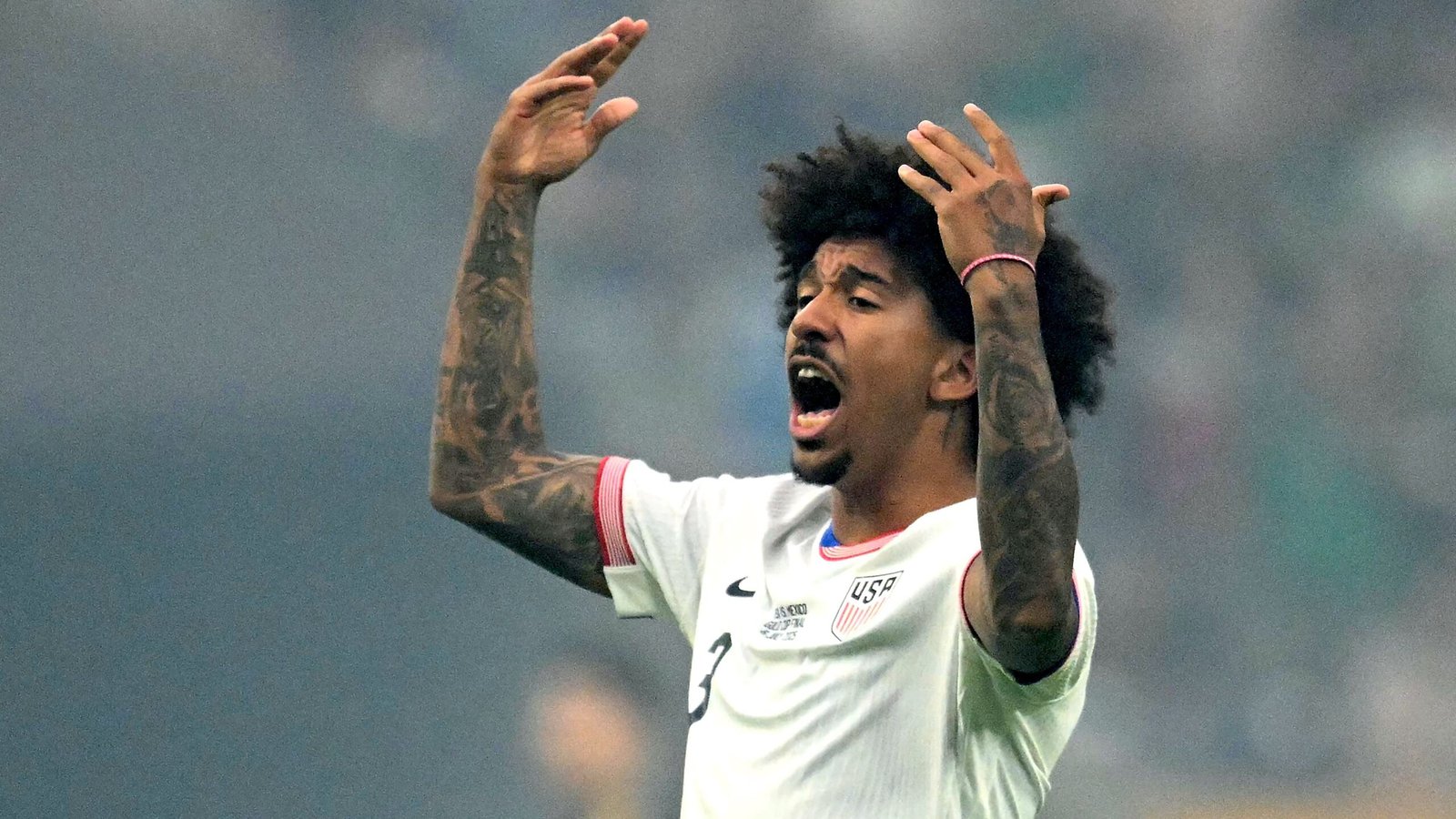 Chris Richards, gekleed in een wit USA-tenue, gebaart gepassioneerd met opgeheven armen en uit frustratie tijdens een wedstrijd.