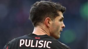Christian Pulisic in een zwart AC Milan-tenue, van achteren gezien, met zijn naam en rugnummer tegen een wazige stadionachtergrond.