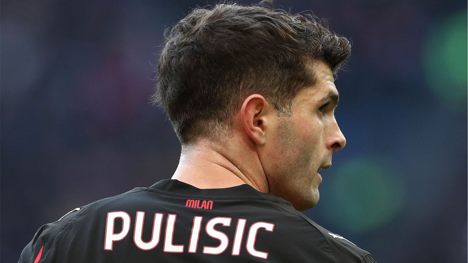 Christian Pulisic in een zwart AC Milan-tenue, van achteren gezien, met zijn naam en rugnummer tegen een wazige stadionachtergrond.
