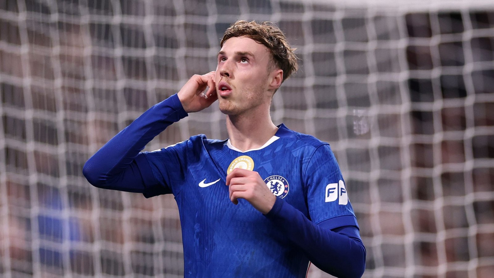كول بالمر من تشيلسي، مرتدياً الزي الأزرق، يرفع نظره متفكراً وهو يمسك بميدالية ذهبية بجانب أذنه، مع وجود شبكة المرمى في الخلفية.