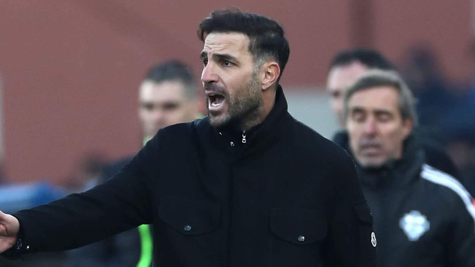 Cesc Fàbregas passionately gestures on the sidelines during a match, wearing a black jacket, with team staff in the background.