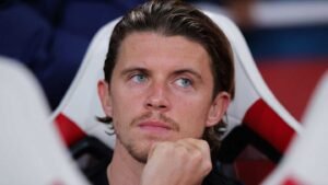 Conor Gallagher sits on the bench with a pensive expression, reflecting intense focus. The Chelsea kit contrasts with the red and white stadium seats.