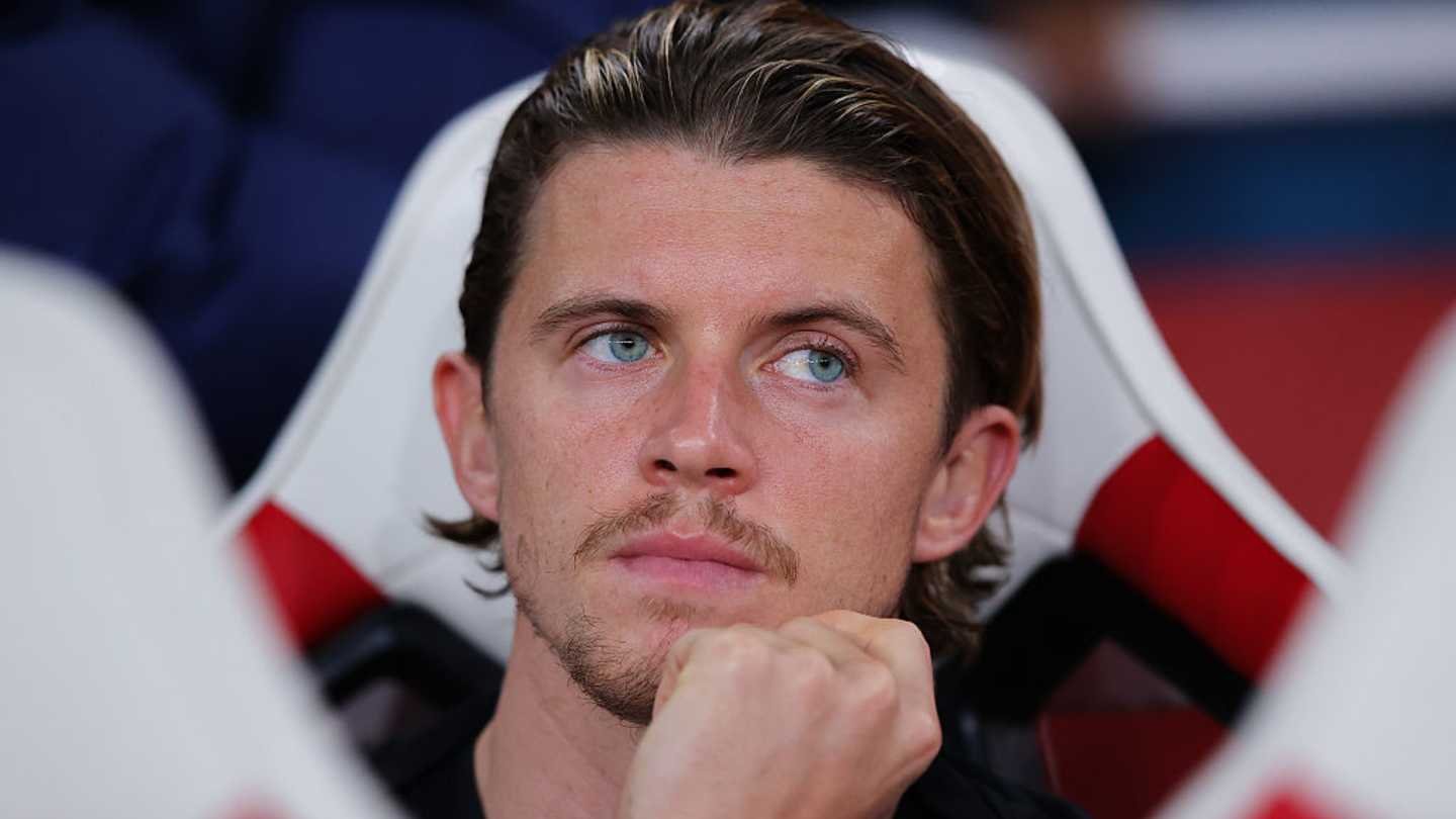 Conor Gallagher sits on the bench with a pensive expression, reflecting intense focus. The Chelsea kit contrasts with the red and white stadium seats.