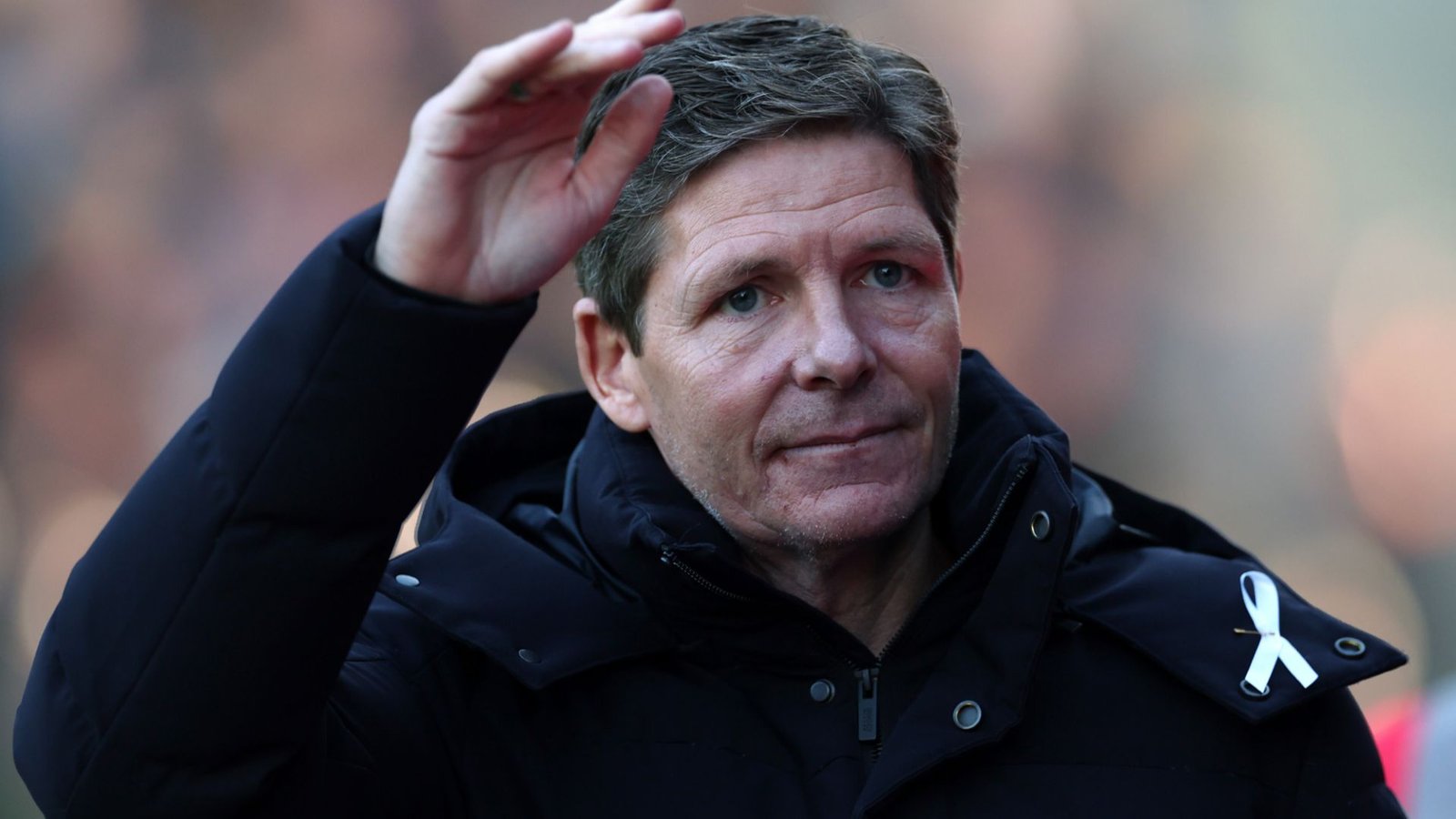 Oliver Glasner zwaait naar het publiek, gekleed in een donkere jas met een wit lint op de schouder gespeld, tijdens een wedstrijdevenement.