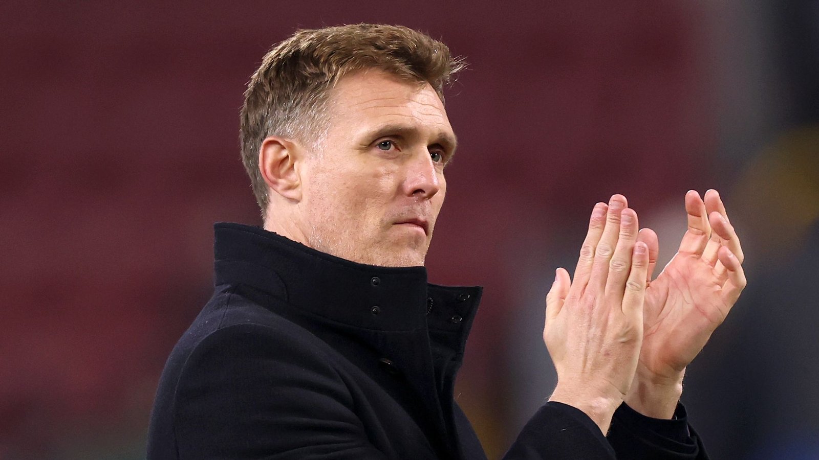 Darren Fletcher, gekleed in een zwarte jas, applaudisseert gepassioneerd, met een wazig stadion op de achtergrond, tijdens een voetbalwedstrijd.