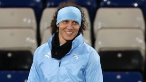 David Luiz lachend in een lichtblauw trainingsjack en hoofdband, staand in een stadion met een vervaagde achtergrond van tribunes.