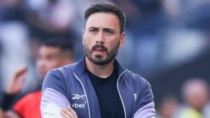 Davide Ancelotti, gekleed in een grijze en paarse trainingsjas, kijkt bedachtzaam vanaf de zijlijn tijdens een voetbalwedstrijd.