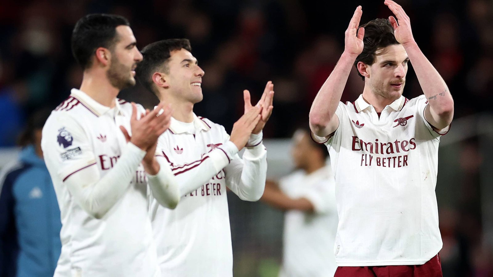 Les joueurs d'Arsenal célèbrent après un match, portant des tenues blanches avec des détails rouges, applaudissant les supporters dans le stade.