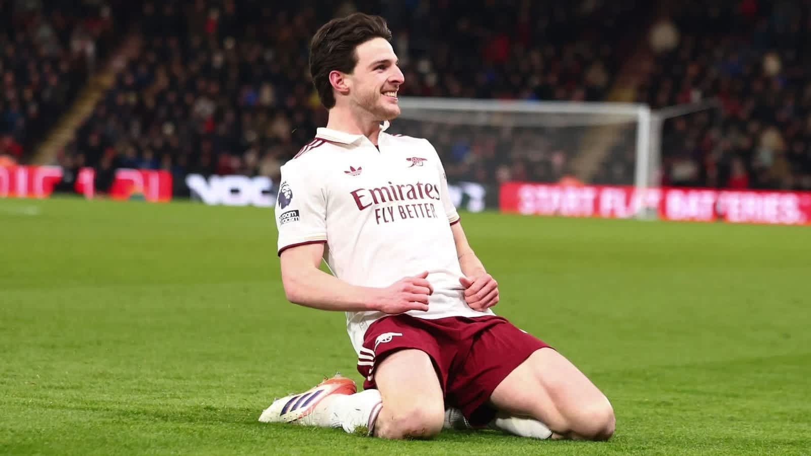 Declan Rice viert op het veld na het scoren voor Arsenal, gekleed in de witte en bordeauxrode outfit van het team, met fans op de achtergrond.