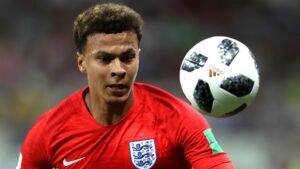 Dele Alli, vêtu du maillot de l'Angleterre, se concentre intensément sur un ballon noir et blanc pendant un match, illustrant une action dynamique.