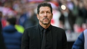 Diego Simeone, in een zwart pak, loopt peinzend voor een wedstrijd, met een vervaagde stadionmenigte op de achtergrond.