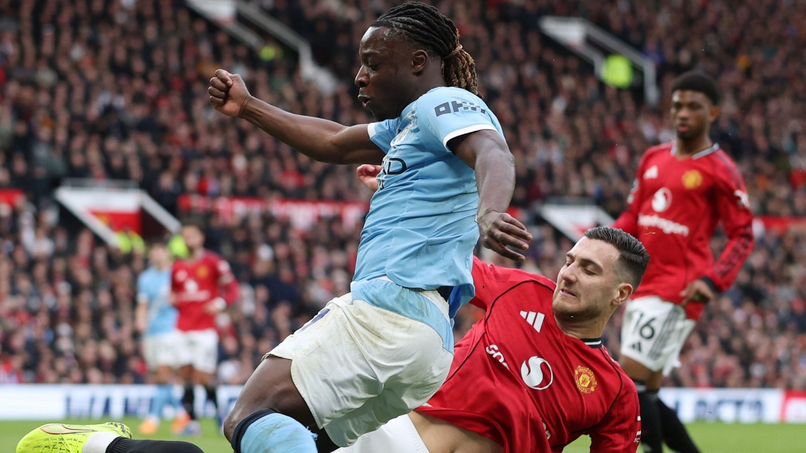 Diogo Dalot challenges Jeremy Doku during a tense Manchester derby, with players in red and blue kit and a packed stadium background.