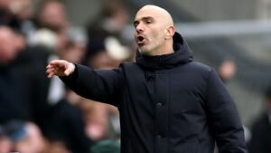 Enzo Maresca gesticule avec passion en bord de terrain, portant un manteau sombre, au milieu d'une foule floue lors d'un match de football.