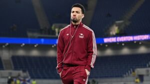 Ethan Nwaneri walking in a maroon Arsenal tracksuit with headphones, set against a stadium backdrop, showcasing a focused expression.