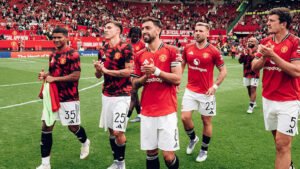 Manchester United players celebrating on the pitch, wearing red and black kits, clapping towards the fans in a vibrant stadium atmosphere.