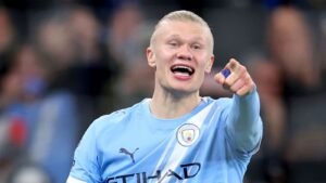 Haaland célèbre dans le ciel bleu de Manchester City, pointant du doigt et souriant joyeusement après un match, affichant son excitation caractéristique.