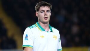 Evan Ferguson staat geconcentreerd op het veld, gekleed in een wit shirt met groene en gele accenten, te midden van een wazige menigte op de achtergrond.