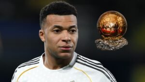 Kylian Mbappé in een wit voetbaltenu, peinzend kijkend, met de Ballon d'Or-trofee prominent naast hem afgebeeld.