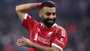 Mohamed Salah glimlachend in een rood Liverpool-tenue tijdens een wedstrijd, waarbij hij zijn kenmerkende baard en kapsel toont.