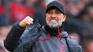 Jurgen Klopp die gepassioneerd viert aan de zijlijn, gekleed in een Liverpool-pet en jas, te midden van een levendige stadionsfeer.
