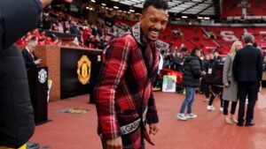 Nani glimlacht terwijl hij een rood geruit gewaad draagt op Old Trafford, met Manchester United-branding zichtbaar op de achtergrond te midden van een uitgelaten menigte.