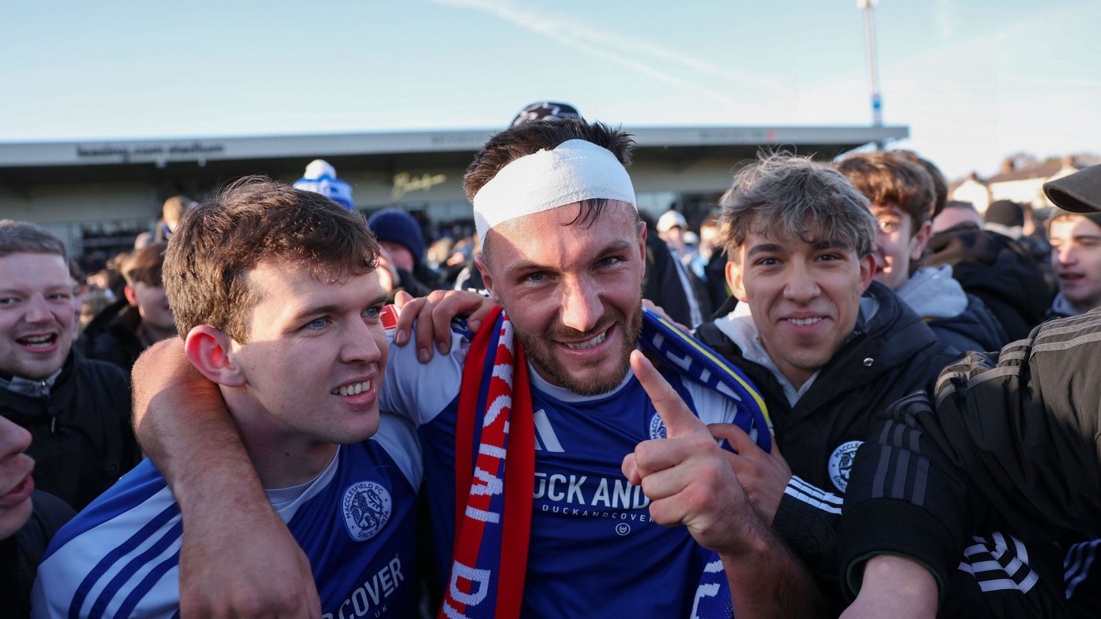 Juichende spelers en fans vieren na een wedstrijd, gekleed in blauwe tenues, waarbij een speler een verband draagt, en stralen vreugde en kameraadschap uit.