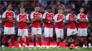 Des joueurs d'Arsenal en tenue domicile rouge sont alignés, applaudissant et concentrés sur un hommage, avec une foule floue en arrière-plan.