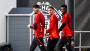 Three Manchester United players in red training kits walk together, smiling, with the club's logo visible in the background.