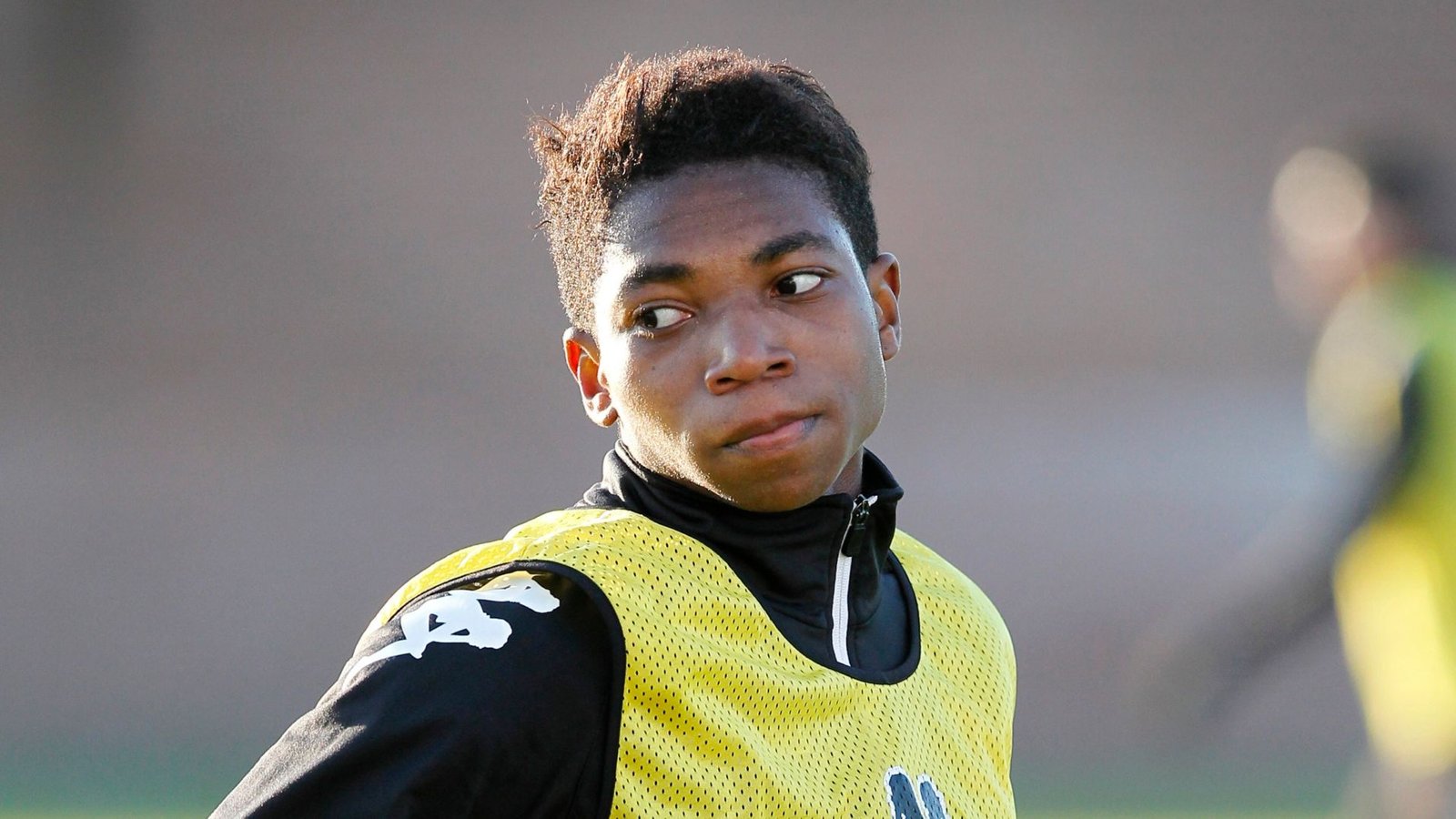 Voormalig Chelsea-jeugdspeler in trainingskleding met een gehesen hesje, peinzend kijkend tijdens een trainingssessie. Vage figuren op de achtergrond.