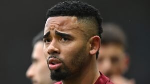 Gabriel Jesus apparaît concentré et déterminé dans une tenue d'entraînement marron, avec des coéquipiers flous en arrière-plan, soulignant son intensité.