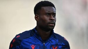 Marc Guehi dans une veste d'entraînement bleue ornée d'accents rouges, concentré et contemplatif, lors d'une séance d'entraînement.