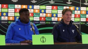 مارك جوهي ومدرب كريستال بالاس يجلسان في مؤتمر صحافي، يرتديان هوديات النادي، مع خلفية تحمل شعارات دوري المؤتمر الأوروبي.