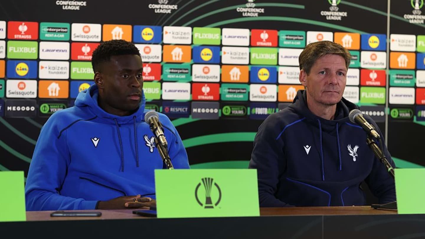 مارك جوهي ومدرب كريستال بالاس يجلسان في مؤتمر صحافي، يرتديان هوديات النادي، مع خلفية تحمل شعارات دوري المؤتمر الأوروبي.