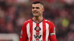 Granit Xhaka lacht terwijl hij het rood-wit gestreepte tenue van Sunderland draagt, met een clubembleem op zijn borst en een wazig stadion op de achtergrond.