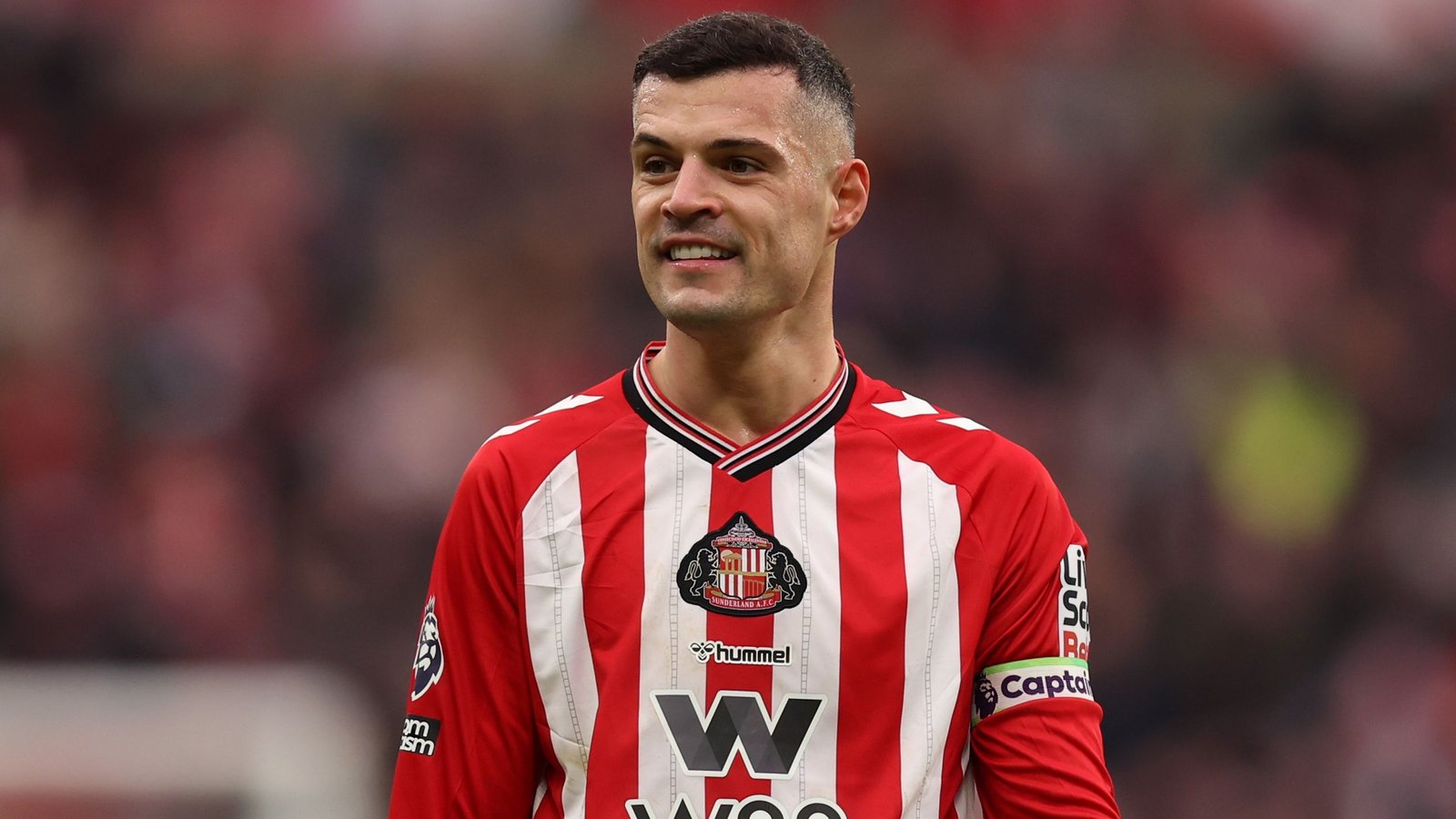 Granit Xhaka lacht terwijl hij het rood-wit gestreepte tenue van Sunderland draagt, met een clubembleem op zijn borst en een wazig stadion op de achtergrond.