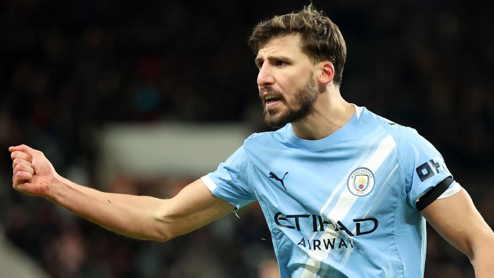 Rúben Dias gesticule avec passion pendant un match, portant le maillot bleu clair de Manchester City avec un brassard de capitaine, illustrant l'esprit d'équipe.