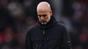 Pep Guardiola avec une expression solennelle, portant une veste sombre, après la défaite de Manchester City lors du derby ; fond de stade flou.