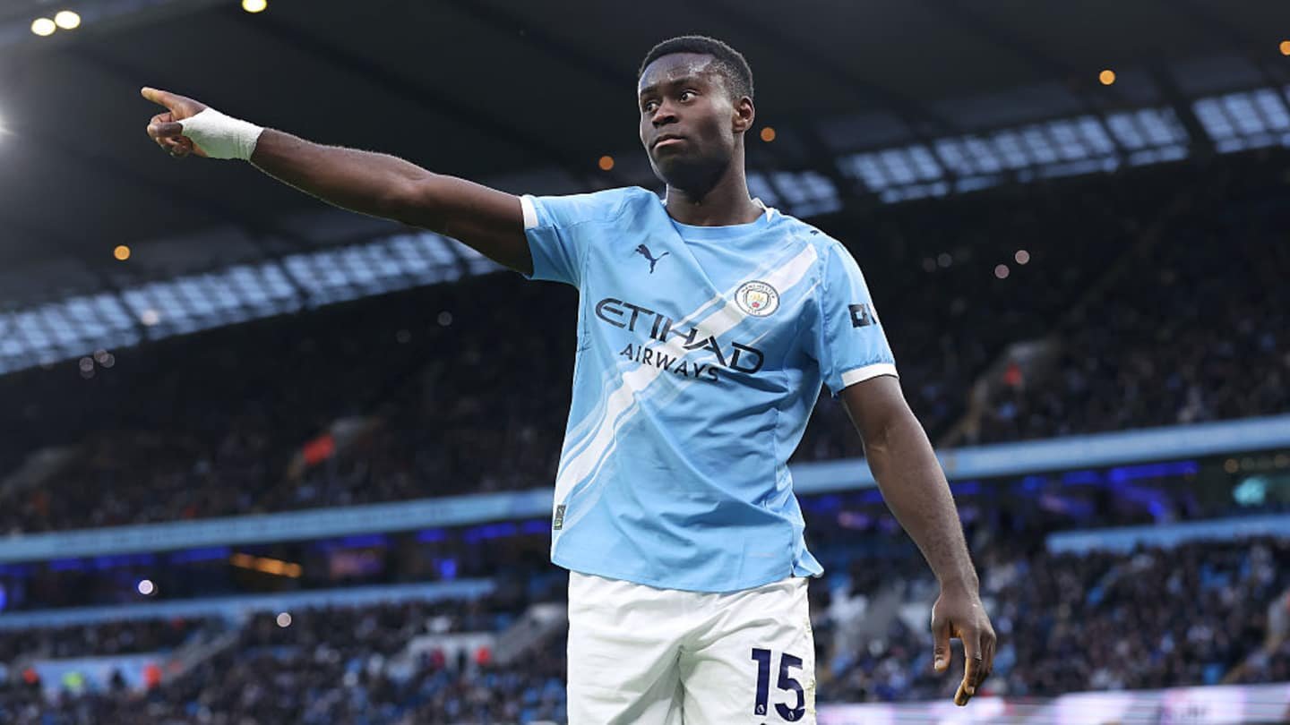 Manchester City player Guehi gestures towards fans, wearing the sky blue home kit with Etihad Airways branding, in a packed stadium.