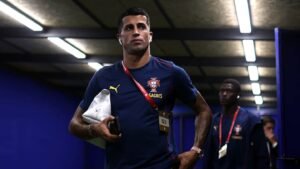 João Cancelo in a blue Portugal training kit walks through a tunnel, holding a phone and earphones, ahead of a match.