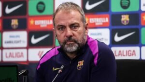 Hansi Flick lors d'une conférence de presse, portant un survêtement du Barça, avec les logos de l'équipe et les marques des sponsors visibles en arrière-plan.