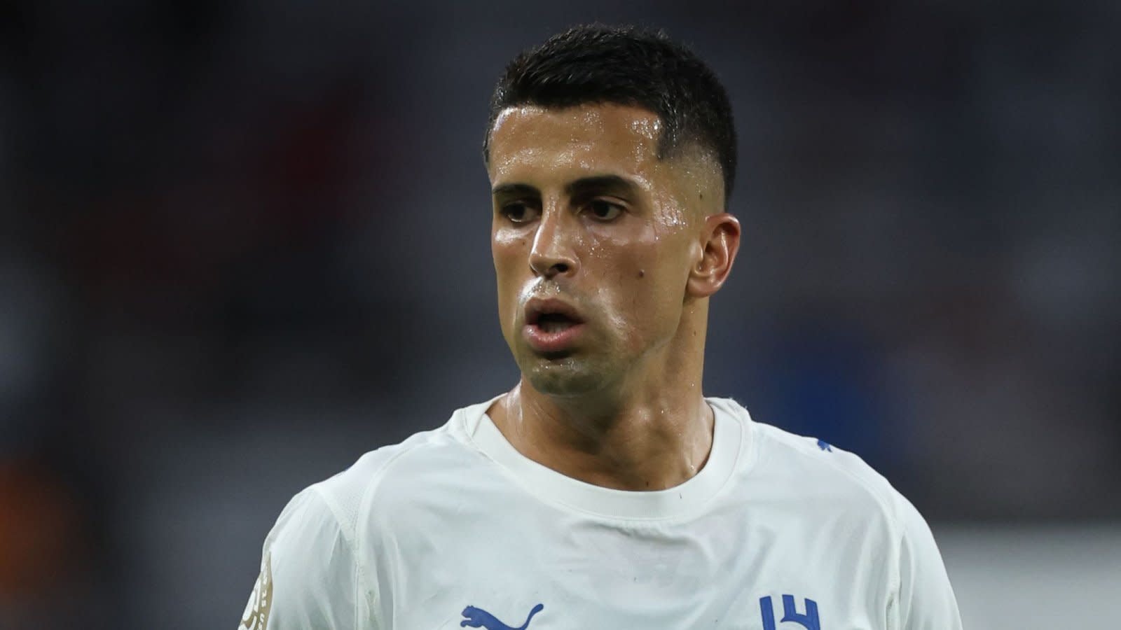 João Cancelo in a white football kit, showing intense focus during a match, with a blurred stadium background capturing the atmosphere.