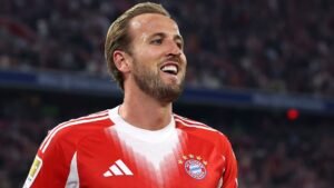 Harry Kane lachend in het rode thuistenue van Bayern München, terwijl hij een doelpunt viert tijdens een Bundesliga-wedstrijd, met fans op de achtergrond.