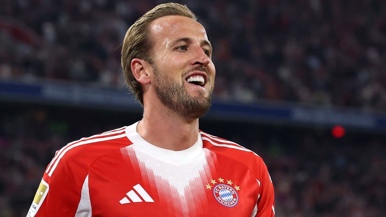 Harry Kane lachend in het rode thuistenue van Bayern München, terwijl hij een doelpunt viert tijdens een Bundesliga-wedstrijd, met fans op de achtergrond.