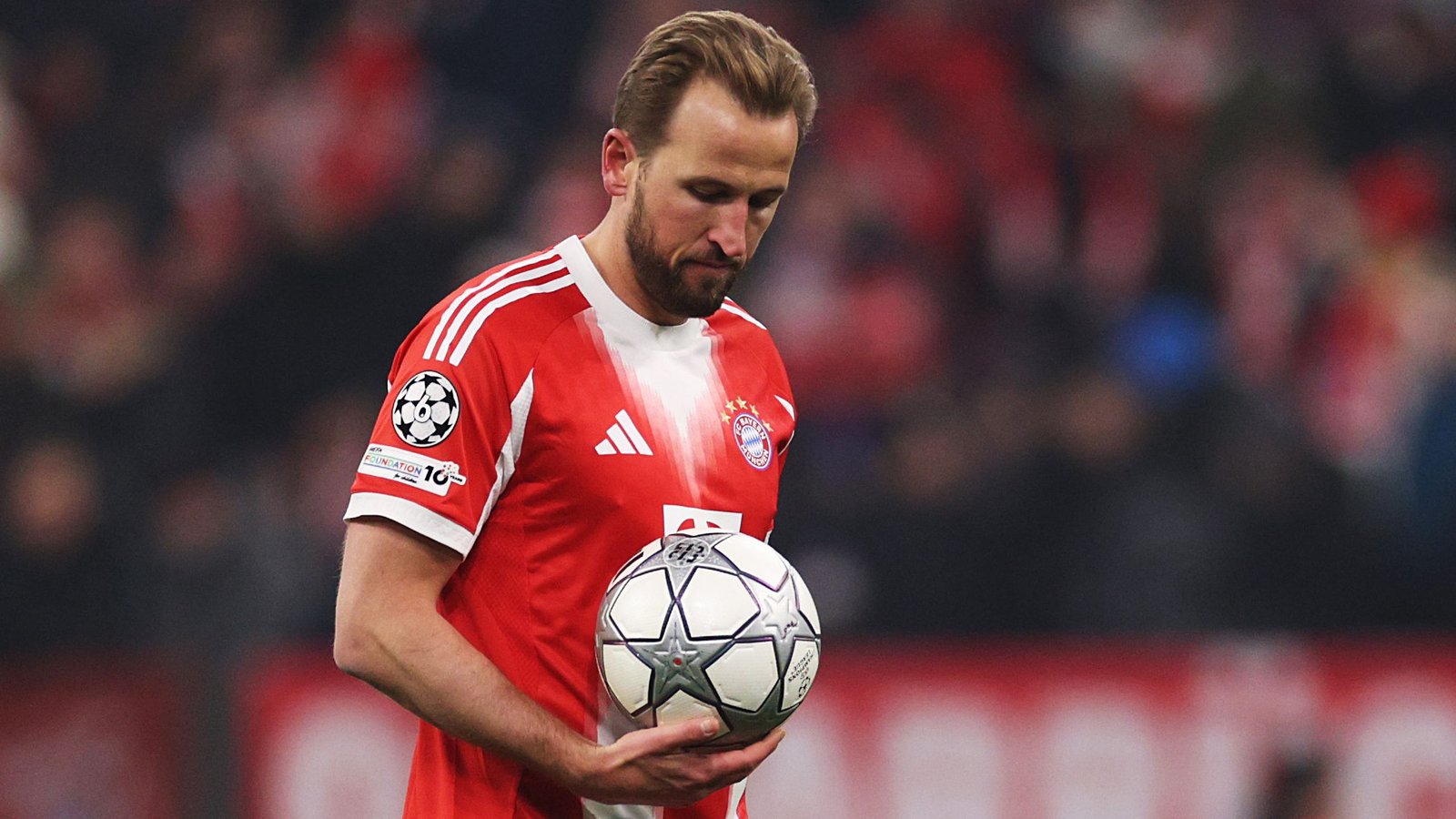 Harry Kane, portant le maillot rouge du Bayern Munich, baisse les yeux vers un ballon de Ligue des Champions, méditant sur un penalty manqué pendant le match.